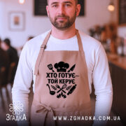 Фартух хто готує керує, бежевий колір, зображення з фронтального ракурсу на чоловікові, фон кафе з розмитими фоновими елементами, принт чорного кольору з кухонними атрибутами.