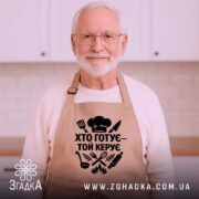 Фартух хто готує керує, бежевий колір, зображення з верхнього ракурсу, фон дерев'яний, принт чорного кольору з кухонними атрибутами.