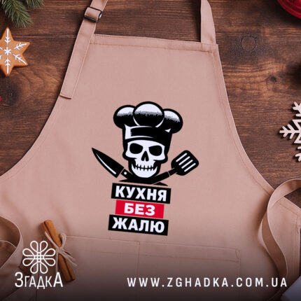 Фартух кухня без жалю, бежевого кольору, зображений на дерев'яному фоні, принт з черепом, кухарським ковпаком та кухонними приладдями, декорований новорічними елементами.