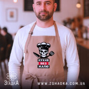 Фартух кухня без жалю, бежевого кольору, зображений на чоловікові в кафе, фон розмитий, принт з черепом, кухарським ковпаком та кухонними приладдями.