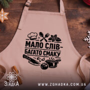 Фартух мало слів, бежевий колір, зображений на дерев'яному фоні, ракурс зверху, принт чорного кольору з написом 'МАЛО СЛІВ - БАГАТО СМАКУ' та кухонними предметами.