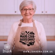 Фартух мало слів, бежевий колір, зображений на манекені з правого ракурсу, фон світлий, принт чорного кольору з написом 'МАЛО СЛІВ - БАГАТО СМАКУ' та кухонними атрибутами.