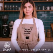 Фартух мама кухня магія, бежевий колір, зображений на жінці в кухні, фон з темно-зеленими плитками, принт з текстом і малюнком казанка.