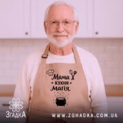 Фартух мама кухня магія, бежевий колір, зображений на дерев'яному фоні з декором, принт з текстом і малюнком казанка.