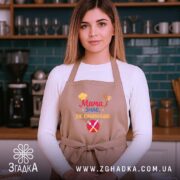Фартух мама знає як бежевий, на жінці в кухонному інтер'єрі, фон темно-зелений, принт з написом 'Мама знає, як смачно' з ілюстраціями кухонного приладдя.