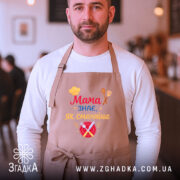 Фартух мама знає як бежевий, на чоловікові в кафе, фон розмитий, принт з написом 'Мама знає, як смачно' з ілюстраціями тарілки і столових приборів.