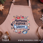 Фартух мамина кухня, бежевого кольору, зображений зверху на дерев'яному столі, фон складається з натурального дерева, принт містить текст "Мамина кухня - закон" та кольорові малюнки овочів і кухонного приладдя.
