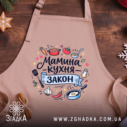Фартух мамина кухня, бежевого кольору, зображений зверху на дерев'яному столі, фон складається з натурального дерева, принт містить текст "Мамина кухня - закон" та кольорові малюнки овочів і кухонного приладдя.