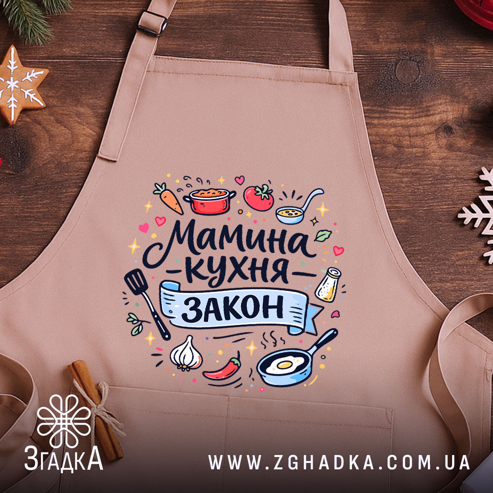 Фартух мамина кухня, бежевого кольору, зображений зверху на дерев'яному столі, фон складається з натурального дерева, принт містить текст "Мамина кухня - закон" та кольорові малюнки овочів і кухонного приладдя.