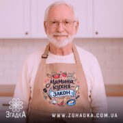 Фартух мамина кухня, бежевого кольору, зображений зверху на дерев'яному столі, фон складається з натурального дерева, принт містить текст "Мамина кухня - закон" та кольорові малюнки овочів і кухонного приладдя.