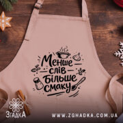 Фартух менше слів більше смаку. Бежевий фартух з чорним принтом, розташований на дерев'яному фоні, деталі принта включають кухонні елементи, такі як каструля, часник і сіль.