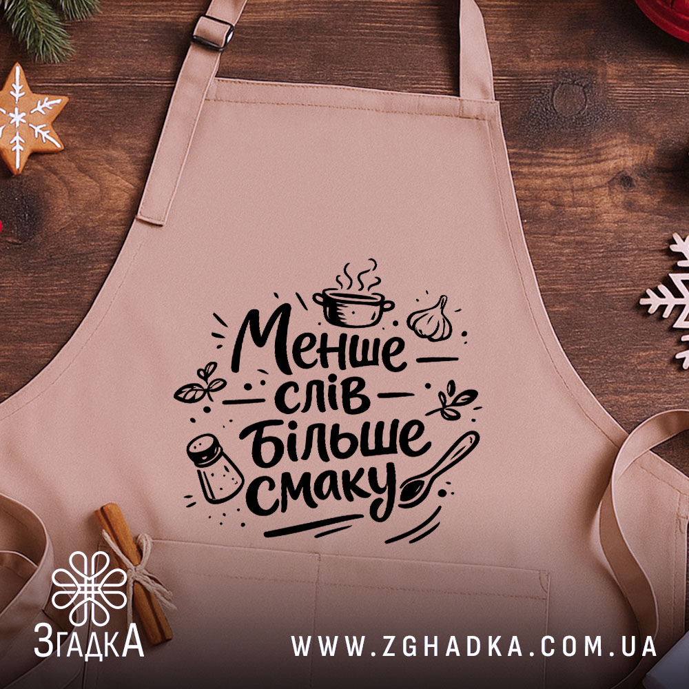 Фартух менше слів більше смаку. Бежевий фартух з чорним принтом, розташований на дерев'яному фоні, деталі принта включають кухонні елементи, такі як каструля, часник і сіль.
