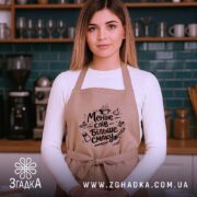 Фартух менше слів більше смаку. Бежевий фартух на жінці, яка стоїть на фоні кухні з темно-зеленими плитками, принт чорного кольору з кухонними атрибутами, жінка має довге волосся.