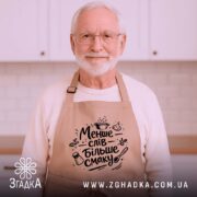 Фартух менше слів більше смаку. Бежевий фартух з чорним принтом, розташований на дерев'яному фоні, деталі принта включають кухонні елементи, такі як каструля, часник і сіль.