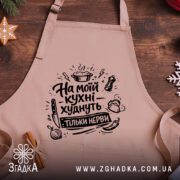 Фартух на моїй кухні, бежевого кольору, з бокового ракурсу, на дерев'яному фоні, принт з написом 'На моїй кухні худнуть тільки нерви' в чорному кольорі, декор у вигляді новорічних елементів.