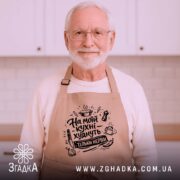 Фартух на моїй кухні, бежевого кольору, з бокового ракурсу, на дерев'яному фоні, принт з написом 'На моїй кухні худнуть тільки нерви' в чорному кольорі, декор у вигляді новорічних елементів.