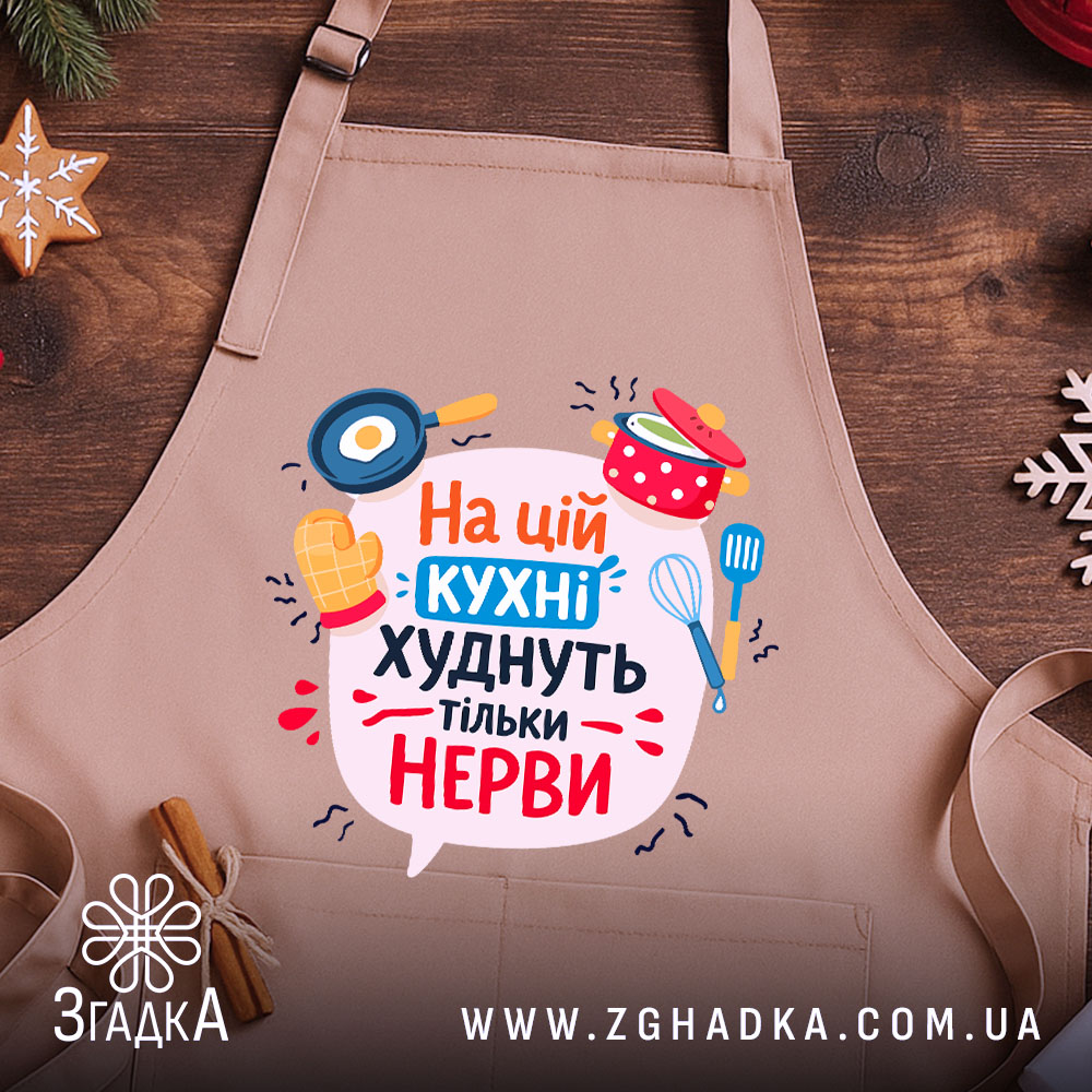 Фартух на цій кухні бежевого кольору, зображений з верхнього ракурсу на дерев'яному фоні, принт містить текст "На цій кухні худнуть тільки нерви" з ілюстраціями кухонного приладдя, поряд розташовані святкові елементи.