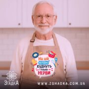 Фартух на цій кухні бежевого кольору, зображений з верхнього ракурсу на дерев'яному фоні, принт містить текст "На цій кухні худнуть тільки нерви" з ілюстраціями кухонного приладдя, поряд розташовані святкові елементи.