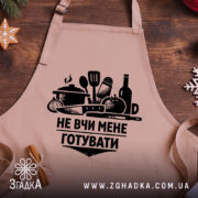 Фартух не вчи мене, бежевого кольору, зображений на дерев'яному фоні, принт чорного кольору з кухонними предметами та написом, на фоні видно новорічні елементи декору.