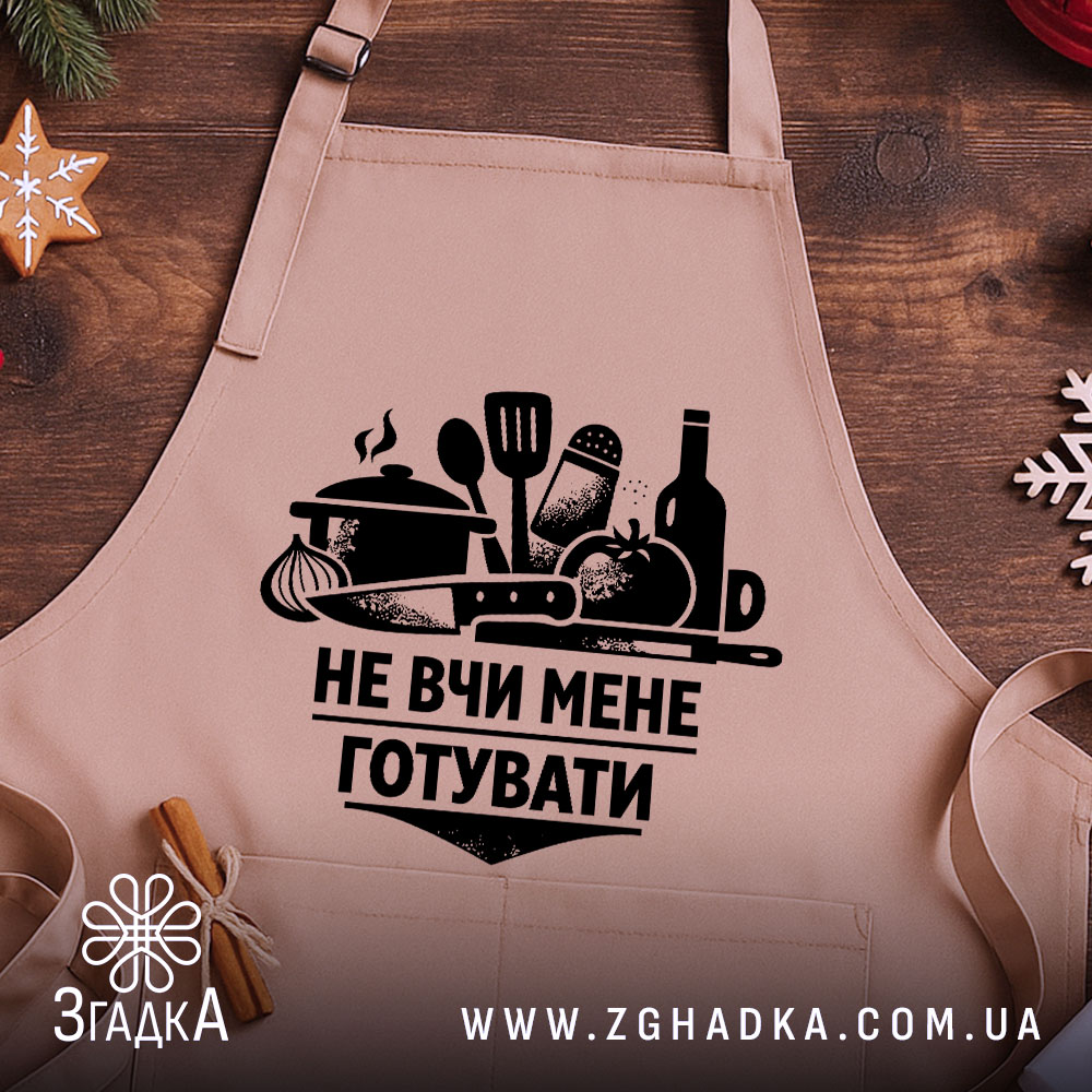 Фартух не вчи мене, бежевого кольору, зображений на дерев'яному фоні, принт чорного кольору з кухонними предметами та написом, на фоні видно новорічні елементи декору.