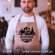 Фартух не вчи мене, бежевого кольору, на чоловікові в кафе, фон розмитий, принт чорного кольору з кухонними предметами та написом.
