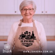 Фартух не вчи мене, бежевого кольору, зображений на манекені, фон світлий, принт чорного кольору з кухонними предметами та написом.