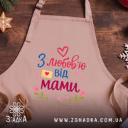 Фартух з любов’ю мами, бежевого кольору, зображений на дерев'яному фоні, принт з написом 'З любов’ю від мами' у яскравих кольорах, поруч з декором у вигляді сніжинки та печива.