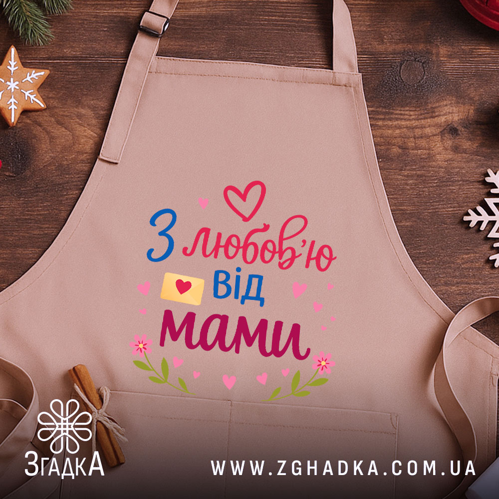 Фартух з любов’ю мами, бежевого кольору, зображений на дерев'яному фоні, принт з написом 'З любов’ю від мами' у яскравих кольорах, поруч з декором у вигляді сніжинки та печива.