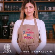 Фартух з любов’ю мами, бежевого кольору, на жінці, фон кухонний з темно-зеленими стінами, принт з написом 'З любов’ю від мами' у яскравих кольорах, жінка усміхається.