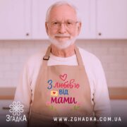 Фартух з любов’ю мами, бежевого кольору, зображений на дерев'яному фоні, принт з написом 'З любов’ю від мами' у яскравих кольорах, поруч з декором у вигляді сніжинки та печива.