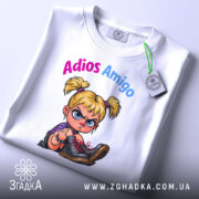 Футболка Adios Amigo білого кольору, зображена з верхнього ракурсу, на ній прикріплена етикетка, фон світлий, принт з малюнком дівчинки з пучками, яка показує середній палець, текст 'Adios Amigo' виконаний у кольорах рожевий та блакитний.