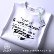 Футболка ангел на мітлі, біла, складена на столі, фон світло-сірий, принт чорного кольору з написом 'Я - ангел, чесне слово! Просто на мітлі ШВИДШЕ.' та етикеткою з зеленим шнурком.