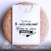 Футболка ангел на мітлі, біла, складена на плетеній основі, фон білий, принт чорного кольору з написом 'Я - ангел, чесне слово! Просто на мітлі ШВИДШЕ.' та видимою етикеткою.