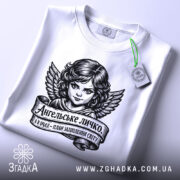 Футболка ангельське личко, біла, складена на столі, фон світло-сірий, принт з зображенням ангела з крилами та написом на стрічці, з етикеткою зеленого кольору.