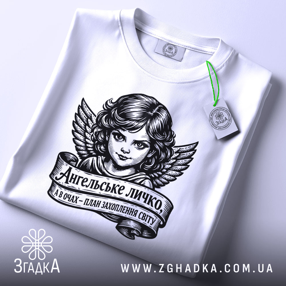 Футболка ангельське личко, біла, складена на столі, фон світло-сірий, принт з зображенням ангела з крилами та написом на стрічці, з етикеткою зеленого кольору.
