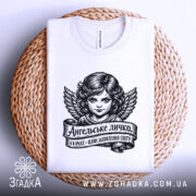 Футболка ангельське личко, біла, складена на плетеній кошику, фон світло-сірий, принт з зображенням ангела з крилами та написом на стрічці.