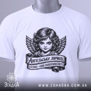 Футболка ангельське личко, біла, складена на столі, фон світло-сірий, принт з зображенням ангела з крилами та написом на стрічці, з етикеткою зеленого кольору.