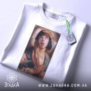 Футболка арт іронія віри, біла футболка, складена на столі, з принтом дівчини з молитвою, фон світло-сірий, на футболці є етикетка з зеленим шнурком.