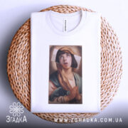 Футболка арт іронія віри, біла футболка, складена на плетеній кошику, з принтом дівчини з молитвою, фон світло-сірий, деталі принта яскраві, видно етикетку на комірці.