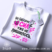 Футболка бабських сил. Біла футболка, складена на столі, з принтом у чорному та рожевому кольорах. На фоні світло-сірого кольору видно етикетку з зеленим шнурком.