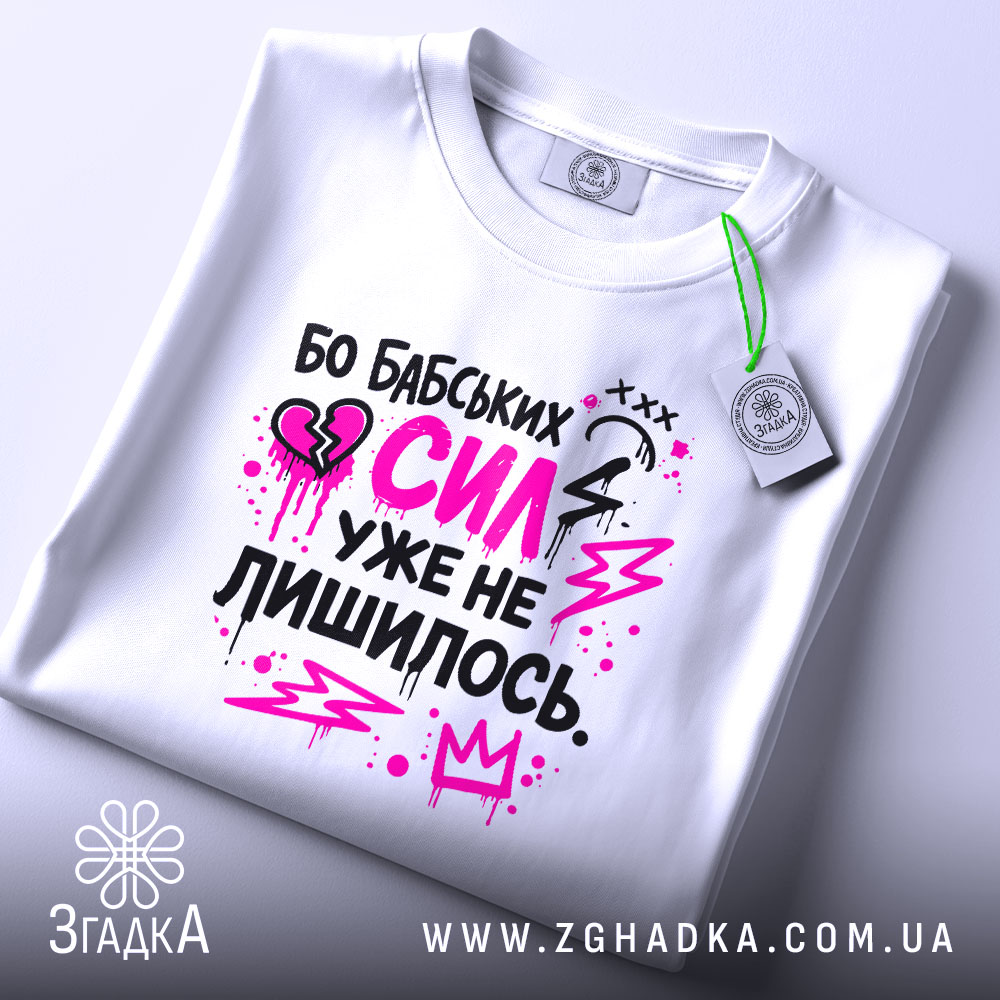Футболка бабських сил. Біла футболка, складена на столі, з принтом у чорному та рожевому кольорах. На фоні світло-сірого кольору видно етикетку з зеленим шнурком.