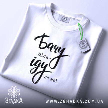 Футболка бачу ціль іду: біла футболка, складена на столі, з видимим етикеткою, фон світло-сірий, принт чорного кольору з написом 'Бачу ціль іду до неї'.