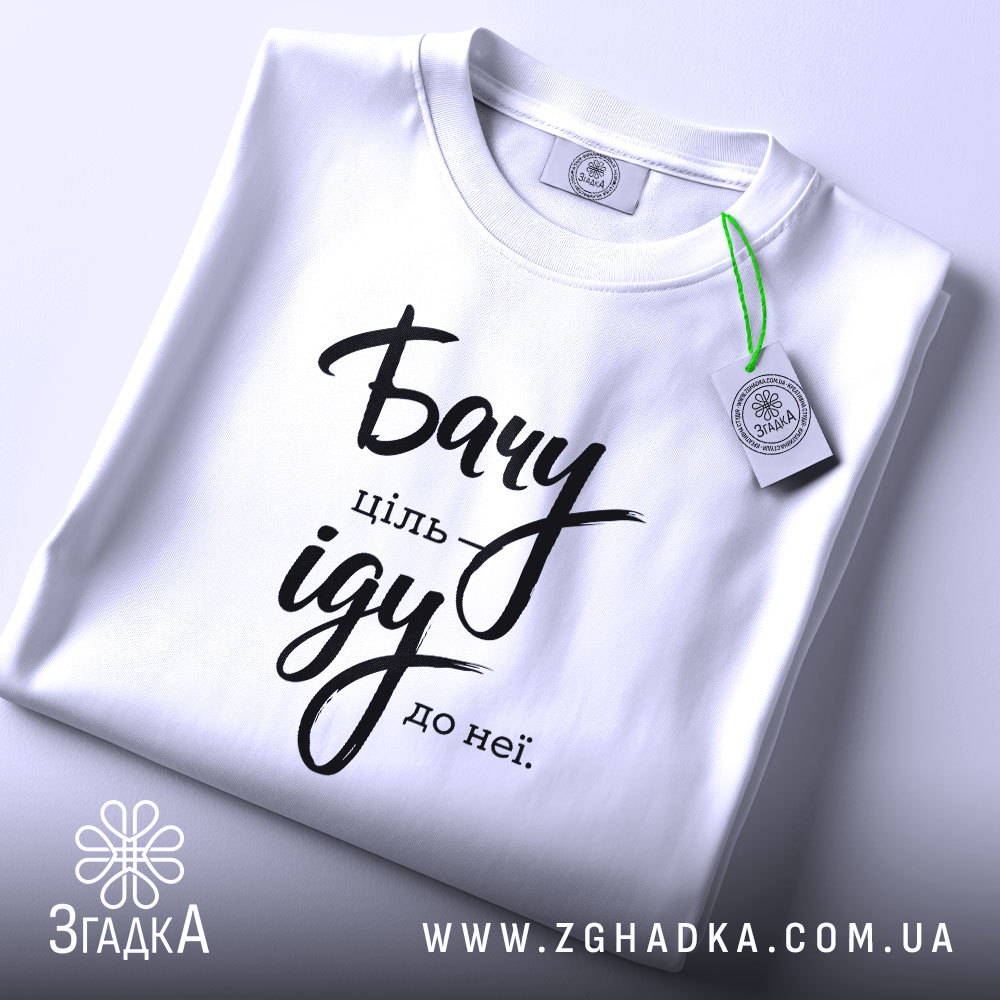 Футболка бачу ціль іду: біла футболка, складена на столі, з видимим етикеткою, фон світло-сірий, принт чорного кольору з написом 'Бачу ціль іду до неї'.