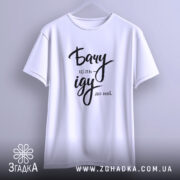 Футболка бачу ціль іду: біла футболка з коротким рукавом, підвішена на вішалці, фон світло-сірий, принт чорного кольору з написом 'Бачу ціль іду до неї'.