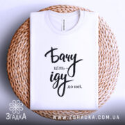 Футболка бачу ціль іду: біла футболка, складена на плетеній кошику, фон світло-сірий, принт чорного кольору з написом 'Бачу ціль іду до неї'.