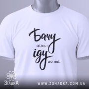 Футболка бачу ціль іду: біла футболка, складена на столі, з видимим етикеткою, фон світло-сірий, принт чорного кольору з написом 'Бачу ціль іду до неї'.