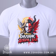 Футболка баланс добра зла, біла, з верхнього ракурсу, на столі, фон світло-сірий, принт з зображенням дівчини з ангельськими і дияволячими елементами, яскраві кольори, червоний, чорний, жовтий, з етикеткою зеленого кольору.