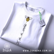 Футболка бджілка це життя. Біла футболка, складена на столі, з видимим принтом бджілки та написом 'Ж.ж.ж.ж.'. На футболці є зелена етикетка, фон світлий, що підкреслює чистоту дизайну.