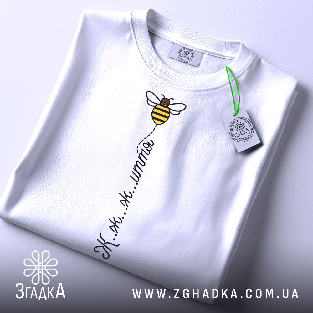 Футболка бджілка це життя. Біла футболка, складена на столі, з видимим принтом бджілки та написом 'Ж.ж.ж.ж.'. На футболці є зелена етикетка, фон світлий, що підкреслює чистоту дизайну.