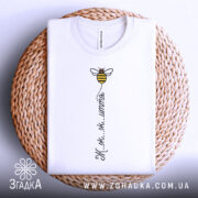 Футболка бджілка це життя. Біла футболка, складена на плетеній кошику, з принтом бджілки та написом 'Ж.ж.ж.ж.'. Фон нейтральний, що підкреслює текстуру кошика.